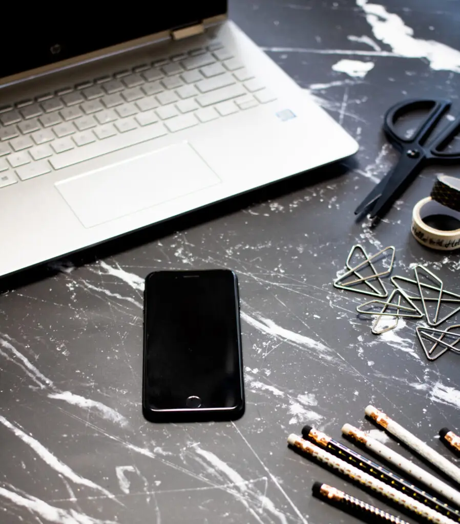 Our Services Image of a desk with an open laptop, iPhone. There is various stationary on the desk (scissors, tape, paperclips and pencils).