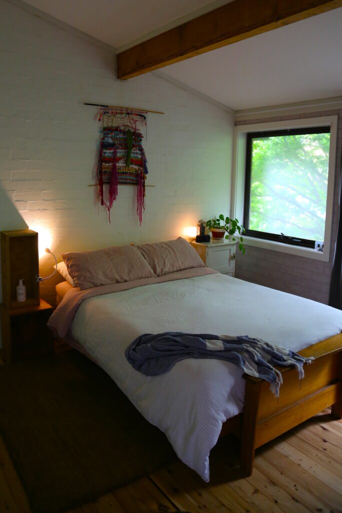 Sydney depicts the naturally lit bedroom in the Sydney property - there is a queen sized bed with light cream linen, peach pillows, and a textile artwork hanging on the wall above the bed