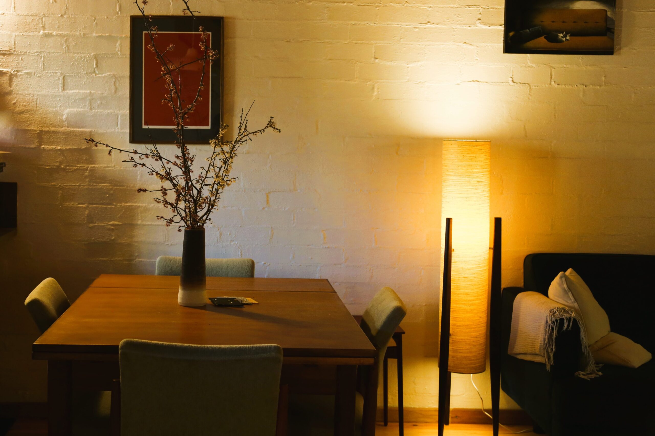 Sydney depicts a timber dining table with four green and timber chairs, a vase with plants, artwork and a warmly litfloor lamp